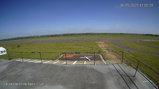A clear view from a webcam overlooking an airfield with a runway in the background and grassy areas on either side. In the foreground, there is a ledge with a metal railing, and below it, a small parking area with designated parking spots. The sky is bright and mostly clear, with a few small clouds. The timestamp in the top right corner reads "16-05-2025 13:00:39," and the bottom left corner displays the text "Leicestershire Aero Club."