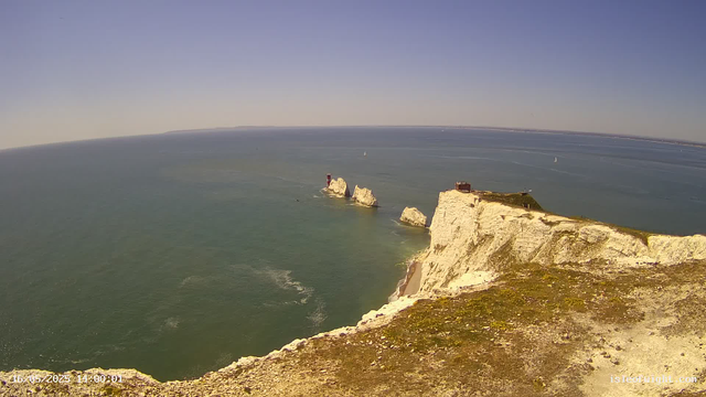 A wide view of white chalk cliffs overlooking the ocean on a clear day. In the distance, there are a few sailboats on the water and a small red lighthouse on a rocky outcrop. The sky is blue, and the water appears calm with gentle waves. The photo is taken from the edge of the cliffs, showing the height and expansive scenery below.
