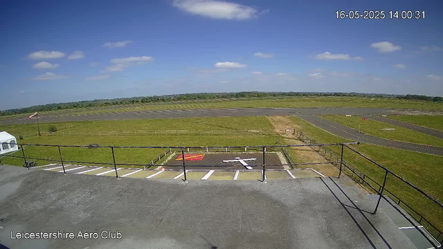 An outdoor view from a webcam showing a grassy area and a runway under a partly cloudy blue sky. In the foreground, there is a small parking area with a few empty parking spaces, a white van on the left side, and a railing along the edge. The date and time stamp in the top right corner reads "16-05-2025 14:00:31," and text in the bottom left corner indicates it is the Leicestershire Aero Club.
