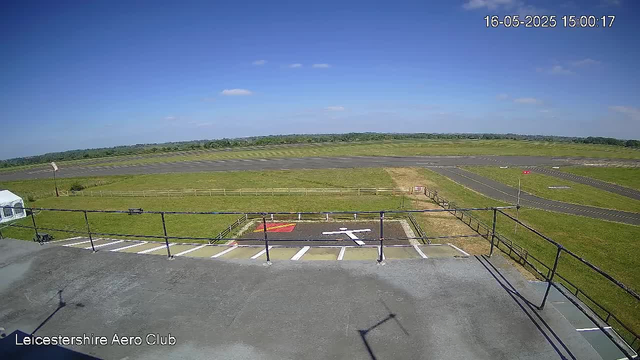 An outdoor view of a small aircraft parking area beside a runway, with a grassy field extending to the horizon under a blue sky with scattered clouds. A white tent is visible on the left, and a fence runs along the perimeter of the parking area. The image has a timestamp in the top right corner showing "16-05-2025 15:00:17," and the bottom left corner displays the text "Leicestershire Aero Club."