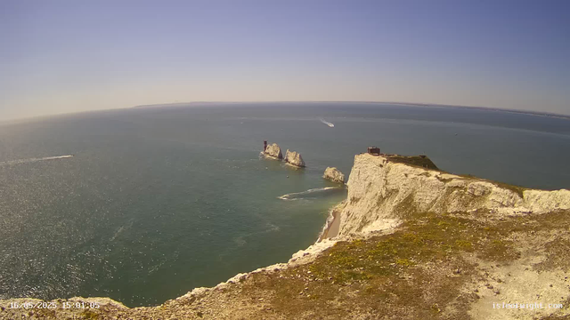 A panoramic view from a high vantage point over the ocean, showing white chalk cliffs with grassy tops extending out to sea. In the water, there are a few large rocks and several boats leaving wakes as they pass by. The sky is clear and blue, and the sunlight reflects off the water's surface.