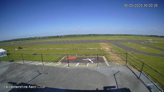 A clear view of an airport runway and taxiways under a blue sky with a few scattered clouds. In the foreground, there is a parking area with marked parking spaces and a helipad with an arrow pointing toward the runway. A small airplane is visible in the distance on the right side of the image. The scene is captured by a webcam at LeicesterShire Aero Club, with the date and time displayed in the top right corner as 16-05-2025, 16:00:26.