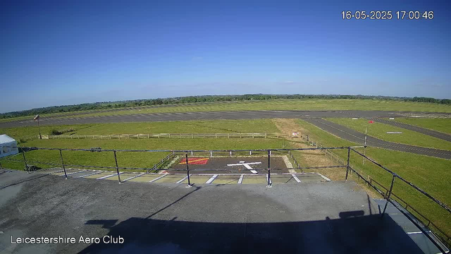A view of an airfield from a webcam, showing a clear blue sky, a grassy area with a small parking space and a helipad marked with a white arrow. There is a fence, a tent on the left, and a paved runway extending into the distance. The image is timestamped "16-05-2025 17:00:46" and labeled "Leicestershire Aero Club" in the bottom left corner.