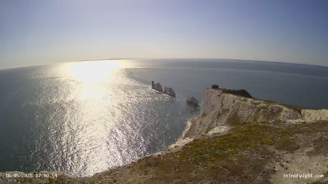 A high-angle view from a webcam shows a rocky coastline with white chalk cliffs overlooking the ocean. The sun is low on the horizon, casting a bright reflection on the water’s surface. A small building and a weathered wooden structure are situated on top of the cliff. In the distance, jagged rocks emerge from the sea, and the sky is clear with a soft gradient from light to darker blue. The timestamp at the bottom left reads "16/05/2025 17:00:54," and the bottom right shows the website "isleofnorth.com."