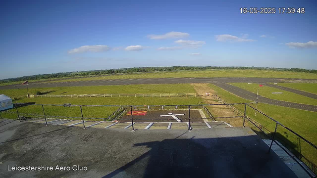 A wide view of an airfield with a grassy area and runways under a blue sky with scattered white clouds. In the foreground, there is a parking area with white markings and a black railing, casting shadows on the surface. The image includes a timestamp in the upper right corner showing "16-05-2025 17:59:48," and the bottom left corner has the text "Leicestershire Aero Club."