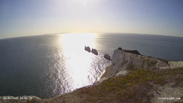 A panoramic view of a rugged coastline during sunset, with white chalk cliffs in the foreground and the ocean stretching to the horizon. The sunlight reflects brightly off the water, creating a shimmering path. In the distance, there are large rock formations emerging from the sea near the coastline.