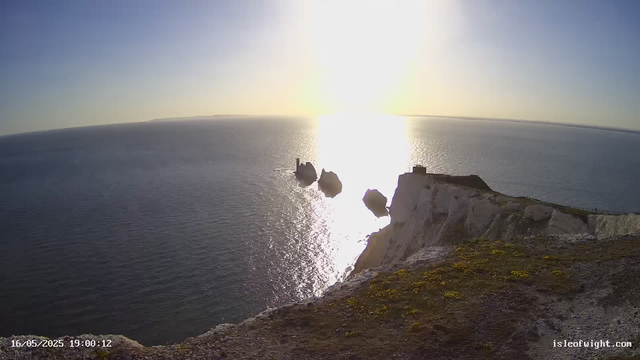 A view from a webcam showing a large body of water with bright sunlight reflecting off its surface. In the distance, there are several large rocks or small islands, with one structure that appears to be a lighthouse. The foreground features a rocky, grassy cliff edge. The sky is clear with a subtle gradient from blue to light near the horizon. The timestamp at the bottom left indicates the date and time as May 16, 2025, at 19:00:12.