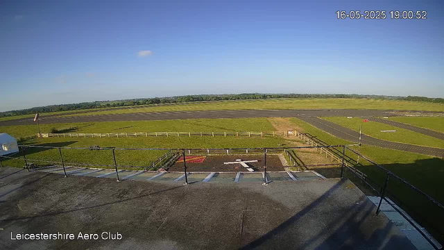 An outdoor view from a camera at Leicestershire Aero Club shows a clear blue sky with a few small clouds. In the foreground, there is a parking area with white lines and a red and white marking. Beyond the parking lot, there is a grassy area with a small fenced-off section. In the background, there are runways or taxiways of an airfield, extending toward the horizon with some markings and a wind sock on the right side. The entire scene is well-lit with sunlight, and the timestamp indicates it was taken on May 16, 2025, at 19:00:52.