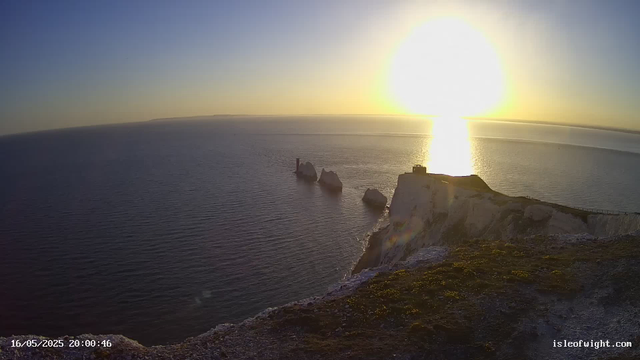A scenic view of the ocean at sunset with white chalk cliffs in the foreground. The sun is near the horizon, casting a bright reflection on the water, and the sky is clear with a gradient from blue to warm tones. There are some rocks and a small structure on the cliff edge. The timestamp shows "16/05/2025 20:00:46," and the website "isleofwight.com" is visible in the corner.