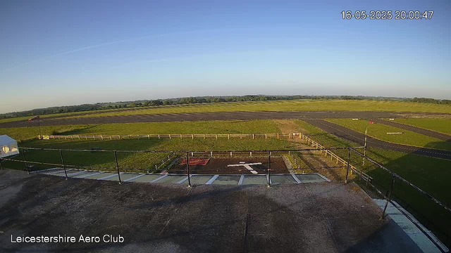An outdoor view from a webcam at Leicestershire Aero Club shows a clear sky with the sun casting long shadows. In the foreground, there is a small paved area with a black railing, a single marked parking spot, and a directional arrow painted on the ground. Beyond the paved area, there is a grassy field enclosed by a beige wooden fence, with a narrow asphalt runway extending into the distance. The landscape consists of open green fields with scattered trees on the horizon under a bright blue sky. The timestamp in the top right corner reads "16-05-2025 20:00:47."