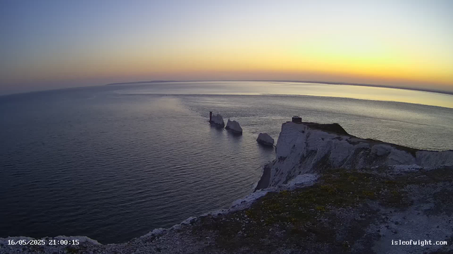 A panoramic view of white chalk cliffs along a coastline at sunset. The sky is a gradient of warm colors, transitioning from yellow to light blue. The ocean below has gentle ripples, and several large rocks are visible in the water near the cliffs. There's a small structure on top of the cliff, and the landscape appears calm and serene. The timestamp on the image reads "16/05/2025 21:00:15," and the website "isleofwight.com" is displayed in the bottom right corner.