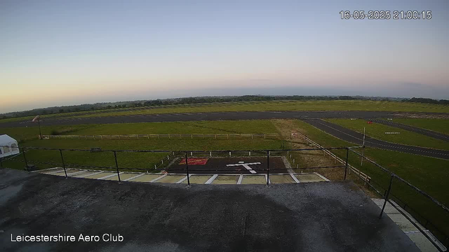 An image of the Leicester Aero Club taken from a webcam shows an open sky during sunset or early evening. The foreground includes a rooftop with a black railing and marked parking spaces, one with a red and white pattern. Beyond the rooftop, there is a paved airfield with taxiways and runways, surrounded by green grass fields and a distant tree line on the horizon. The date and time in the top right corner read "16-05-2025 21:00:15."