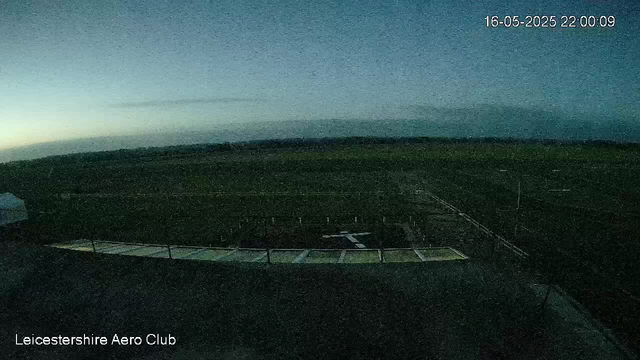 A dark, nighttime view from a webcam shows a large open field with a structure that appears to be a rectangular platform or stage in the foreground. The sky is mostly clear with a subtle gradient from light to dark. The top right corner displays the date and time: 16-05-2025 22:00:09. The bottom left corner contains the text “Leicestershire Aero Club.”