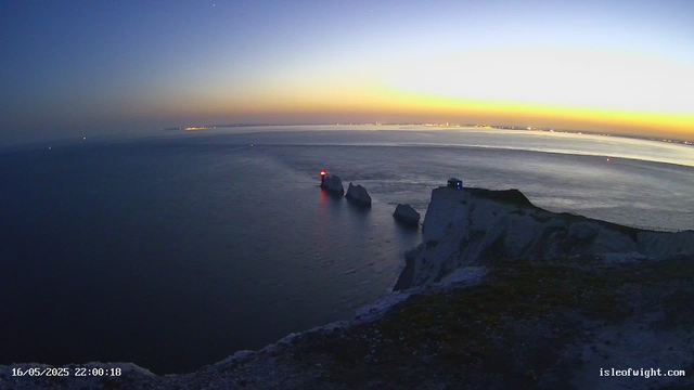 A view of a coastal landscape during dusk or dawn, featuring white chalk cliffs with a grassy top, two small lighthouses on the cliff edge (one with a red light, another with a blue light), and several large rocks protruding from the water below. The sky is a gradient of deep blue to light yellow near the horizon, with the water calm and reflecting the colors of the sky. In the distance, there are faint lights from a town or city along the horizon. The timestamp and website are visible at the bottom of the image.