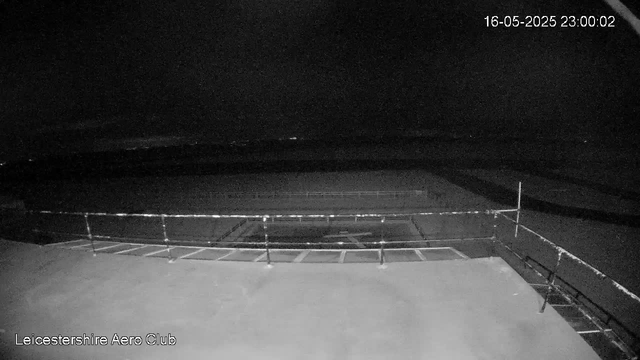 A black-and-white image from a webcam showing an empty outdoor area at night. The scene includes a fenced enclosure in the foreground with parking spaces visible, and a dark, open space beyond. The top right corner displays the date and time as 16-05-2025 23:00:02. The bottom left corner has the text "Leicestershire Aero Club."