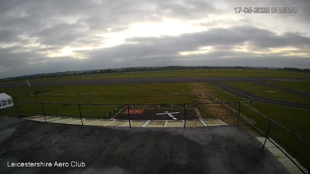 A view from a webcam at Leicestershire Aero Club shows an overcast sky with the sun trying to break through gray clouds. The scene includes a grassy airfield with a runway and taxiways stretching into the distance. In the foreground, there is a terrace or observation deck with a black metal railing. The timestamp in the upper right corner indicates the date and time as May 17, 2025, at 4:59 AM. The image has some empty parking spots and a small flagpole on the left side.