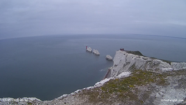 A view from atop white chalk cliffs overlooking a calm sea with large rock formations jutting out of the water and a lighthouse visible on the far-left rock. The sky is overcast with gray clouds.