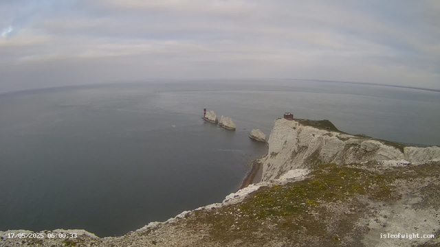 A view from a webcam showing white chalk cliffs extending into the sea with a small lighthouse or marker on one of the cliffs. The water is calm, and the sky is partly cloudy with patches of blue. There is a timestamp at the bottom left corner indicating "17/05/2025 06:00:33," and a website watermark in the bottom right corner reading "isleofwight.com."