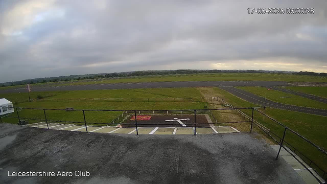 An outdoor view of an airfield under cloudy skies, with a section of a parking area and a small helipad in the foreground, and a runway extending into the distance across green fields. The image is timestamped "17-05-2025 06:00:28" in the upper right corner, and "Leicestershire Aero Club" is written in the bottom left corner.
