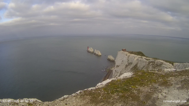 An aerial view of white chalk cliffs with three large rock stacks extending into the sea. There is a lighthouse on one of the stacks and a small building on the cliff's edge. The sky is partly cloudy with patches of sunlight. The date and time displayed are 17/05/2025 at 07:00:42.
