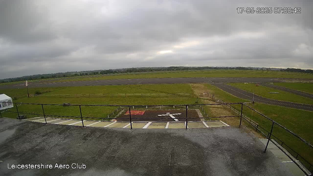 An empty parking area with a railing in the foreground, overlooking a grassy field and a racetrack. Overcast gray clouds cover the sky, and a timestamp in the upper right corner reads "17-05-2025 07:00:42." The bottom left corner has a label that says "Leicestershire Aero Club."