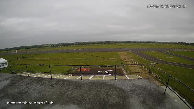 A view from a webcam at Leicesterhire Aero Club showing a cloudy sky over a runway and grassy fields. The image includes a parking area with white lines and a red marking. The scene appears quiet with no people or aircraft visible. The timestamp in the upper right corner reads "17-05-2025 08:00:14."