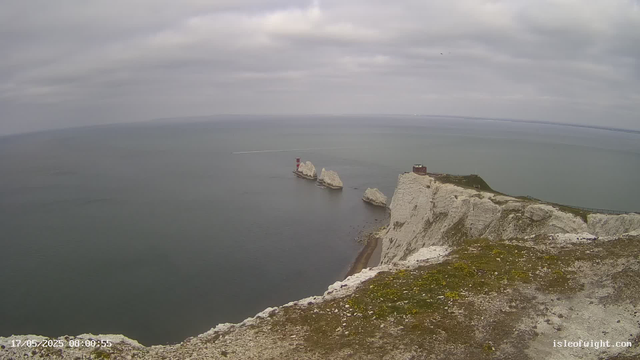 A view of the white chalk cliffs extending into the sea with a small building and path on the cliff top. Two prominent rock stacks are visible offshore, and a lighthouse is situated on one of the stacks. The sky is overcast with gray clouds. The water below is calm, and a boat's wake is visible in the distance. The image is timestamped "17/05/2025 08:00:55" and has a website URL "isleofwight.com" at the bottom right.