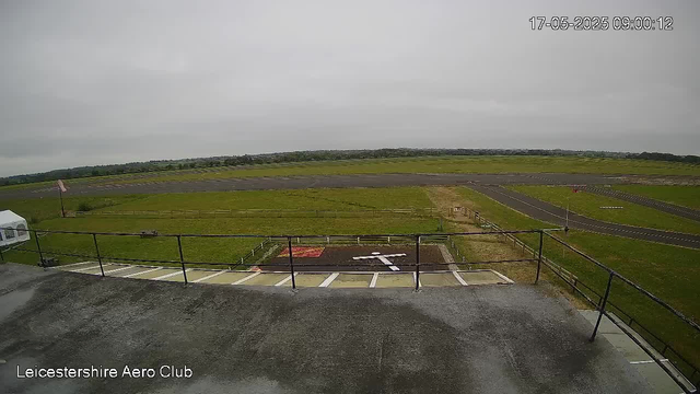 An overcast view of a small airfield with a runway stretching into the distance. In the foreground, there is a balcony or observation area with a metal railing. To the left, part of a white tent or canopy is visible. The image includes a timestamp of 17-05-2025 09:00:12 and the label "Leicestershire Aero Club" at the bottom left corner. The sky is cloudy, and the surrounding landscape is mostly flat with some distant trees.