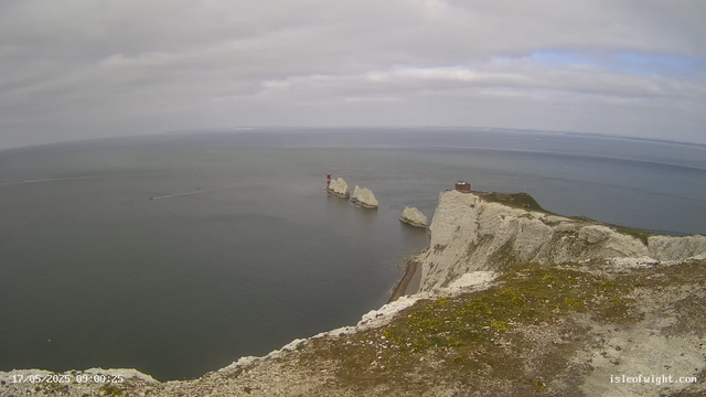 A daytime view from a high vantage point overlooking white chalk cliffs extending into the sea. There are three large rocks or sea stacks near the water's edge, with a small red and white lighthouse atop one of the rocks. The ocean appears calm, with boats leaving white trails on the water. The sky is mostly cloudy with patches of blue, and the horizon stretches far into the distance. The bottom left corner shows the date and time, "17/05/2025 09:00:25," and the bottom right has a website URL, "isleofwight.com."