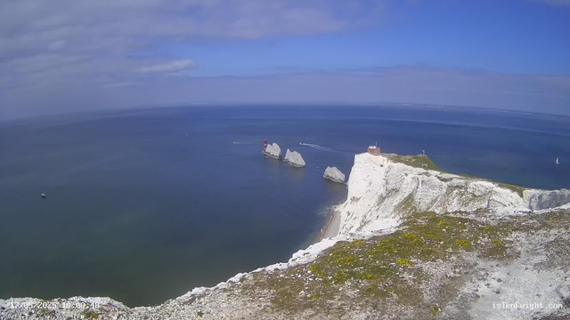 A panoramic view of white chalk cliffs overlooking the ocean, with a lighthouse and a small building situated on top of the cliffs. There are large rock formations in the water near the base of the cliffs, and a sailboat and a small boat are visible in the distance on the sea. The sky is partly cloudy with patches of blue.