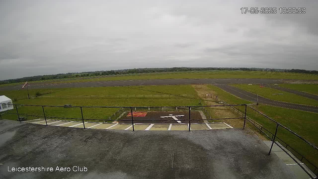 A view from a webcam at Leicesterhire Aero Club showing an overcast sky with grey clouds. The scene includes a runway and green grassy areas surrounding it, with some empty parking spaces in the foreground. The timestamp in the top right corner indicates the date as May 17, 2025, at 10:00:59.