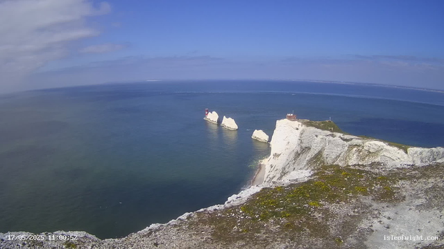 Aerial view of white chalk cliffs with patches of green grass, overlooking a blue ocean with rock stacks and a lighthouse on top of one of the cliffs. The sky is mostly clear with some scattered clouds. The image is timestamped "17/05/2025 11:00:52" at the bottom left corner.