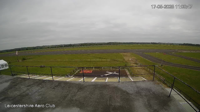 A view from a webcam showing an open space with a grassy field, a runway, and a cloudy sky. In the foreground, there is a concrete area with a railing and a small parking area. The timestamp in the top right corner reads "17-05-2025 11:00:47." The bottom left corner displays the text "Leicestershire Aero Club."