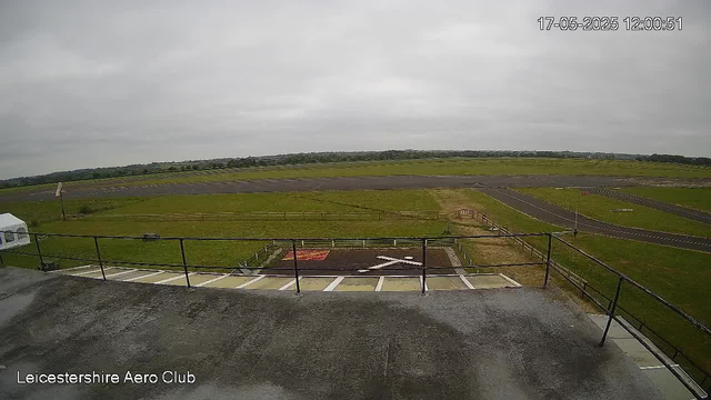 A view from a webcam at Leicestershire Aero Club shows an empty parking area and a runway in the distance under a cloudy sky. There are green grassy fields surrounding the runway, with no aircraft or people visible. The timestamp in the top right corner reads "17-05-2025 12:00:51."