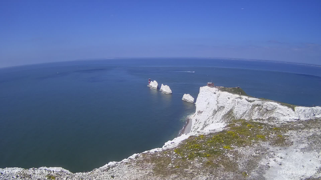 A stunning view of white chalk cliffs with a lighthouse and a small building on top, overlooking a calm blue sea under a clear blue sky. The cliffs extend to the right, with the water stretching out to the horizon. There are a few boats flying across the water, leaving white trails behind them. The scene appears to be bright and sunny.