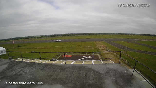 An overcast view of an airfield taken from a webcam. In the foreground, there is a balcony with a metal railing, some parking spaces, and a small marked area on the ground. Beyond the balcony, there are grassy areas and taxiways. In the background, a small airplane is parked on the tarmac. The sky is gray with thick cloud cover, and the timestamp in the top right corner reads "17-05-2025 13:00:47". The bottom left corner has text that says "Leicestershire Aero Club."