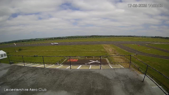 A view from a webcam overlooking an airfield with a small airplane taxiing on the runway in the distance. The sky is partly cloudy with some patches of blue, and the landscape is mostly flat with grassy areas. There is a parking area in the foreground with white parking lines and a black railing along the edge. The image is timestamped "17-05-2025 14:00:03" and has a label at the bottom left that reads "Leicestershire Aero Club".