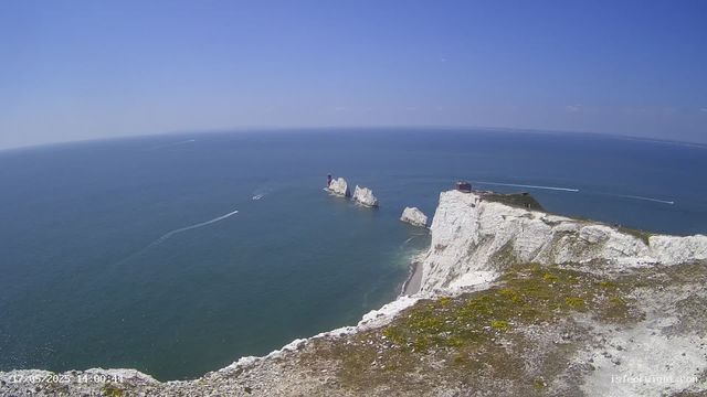 A wide aerial view of white chalk cliffs overlooking the sea on a clear, sunny day. Several boats are sailing in the water, leaving white trails behind them. The sky is bright blue with a few wispy clouds. At the top of the cliffs, there is a small building or structure. The foreground shows the rocky edge of the cliff with some green moss or grass. The timestamp on the bottom left indicates the date and time as May 20, 2023, at 14:00:44.