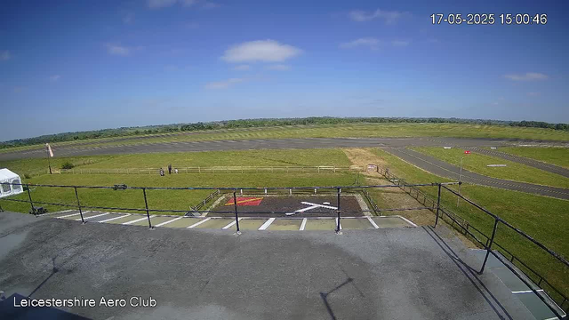 An outdoor scene at Leicester Shire Aero Club featuring a clear blue sky with scattered clouds. In the foreground, there is a safety railing and a small helicopter landing pad with a red border and a white helicopter symbol. To the left, a white tent is visible. Beyond the landing area, there is a grassy field with a few people standing near a wooden fence. In the background, a paved runway extends across the scene, with a few small structures or markers along its length. The date and time displayed on the top right corner are 17-05-2025 at 15:00:46.