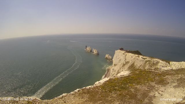 A view from a high cliff overlooking the ocean, with white chalk cliffs extending into the sea. There are three prominent rock stacks in the water near the base of the cliffs. The sky is clear with a few small clouds near the horizon, and boats are visible moving across the water, leaving white trails behind them. The photo was taken in the late afternoon, as indicated by the timestamp.