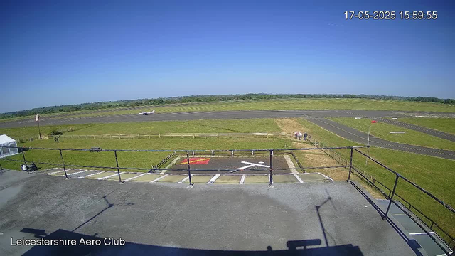 A view from a webcam at Leicestershire Aero Club showing a sunny day at an airport. In the background, a small aircraft is taxiing on the runway, and the surrounding landscape features green fields and a clear blue sky. The image is timestamped "17-05-2025 15:59:55".