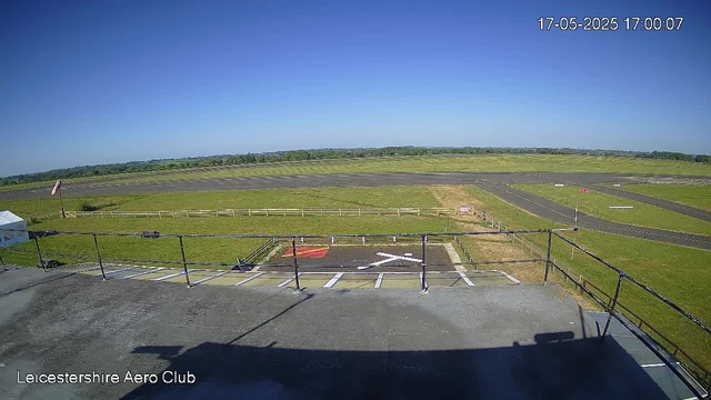 A view from a webcam at Leicestershire Aero Club showing an empty tarmac area with parking spaces and a small helipad in the foreground. Beyond the tarmac, there are grassy fields and a runway extending into the distance under a clear blue sky. The scene appears calm and sunny, with no aircraft or people visible. The timestamp in the top right corner indicates the date and time as 17-05-2025 at 17:00:07.