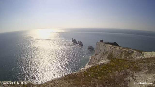 A panoramic view of a coastal landscape during sunset, featuring high white chalk cliffs with patches of green vegetation on top. In the distance, several large sea stacks are visible rising from the ocean, with one having a distinct tower-like structure. The sunlight reflects off the ocean's surface, creating a shimmering effect. The sky is clear with a pale blue hue. The foreground shows the edge of the cliff with some moss or grass growing on it.