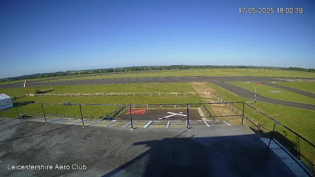 A clear, sunny day at Leicestershire Aero Club with a wide view of an airfield. In the foreground, there is a black asphalt rooftop with a railing, and below that, a concrete area with parking spots and white painted markings. Further back, the grassy airfield is bordered by a low wooden fence, with a small runway marked on the grass. The background features open fields and a distant tree line under a bright blue sky. The timestamp reads 17-05-2025 18:00:39.