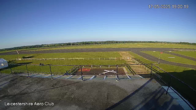 A wide view of an airfield with grassy runways under a clear blue sky. The photo is taken from a high vantage point, showing the tarmac, parking area, and surrounding open fields. There is a railing in the foreground, and the image includes a timestamp in the upper right corner and the text "Leicestershire Aero Club" at the bottom left corner.