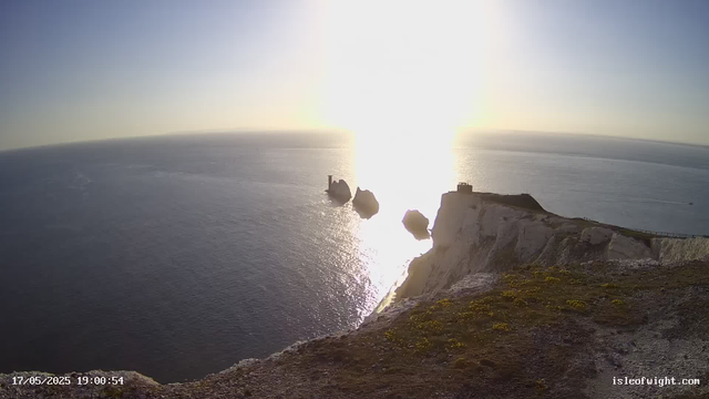 A view of a coastal landscape at sunset, with the sun low on the horizon casting a bright reflection on the ocean. White chalk cliffs with patches of grass and moss are visible in the foreground, and four separate sea stacks rise from the water, with a structure on top of the tallest one. The scene is peaceful and illuminated by warm sunlight. The timestamp shows the date and time as 17/05/2025 at 19:00:54.