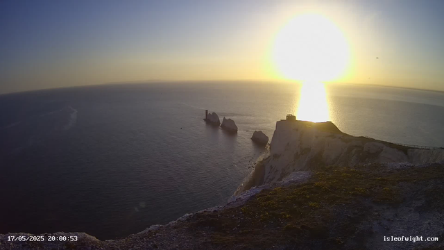 A view of white chalk cliffs overlooking the ocean at sunset, with the sun near the horizon casting a bright reflection across the water. Rock formations and a small building or structure are visible on the cliff edge. The sky is mostly clear with a few birds flying. The timestamp indicates the date and time as May 17, 2025, at 8:00:53 PM.