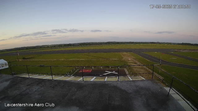 An empty airfield runway with a small helipad in the foreground. The sky is mostly clear with a few clouds, and the landscape stretches out into the distance with grassy areas and a paved taxiway. The image has a timestamp showing May 17, 2025, at 9:00 PM and a label indicating it is from the Leicester-Shire Aero Club.