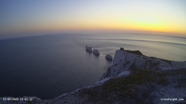 A view of the white chalk cliffs of Étretat at sunset, with the sea stretching out below. The sky has a gradient of soft colors, transitioning from pale yellow near the horizon to a light blue higher up. Three large rocks are visible in the water near the cliffs, and a small building is situated on the edge of the cliff. The foreground shows the rocky, grassy terrain of the cliff's edge. The date and time are displayed at the bottom left corner.