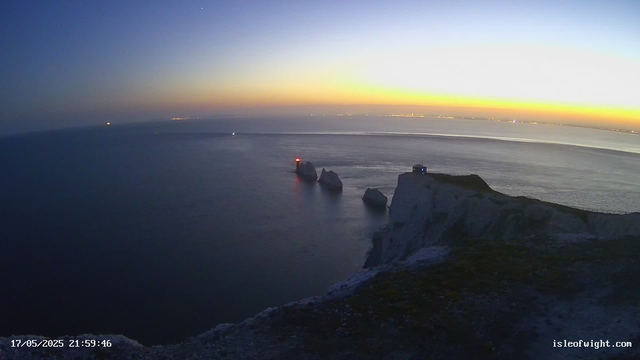 An ocean view at dusk with a sky transitioning from orange to dark blue. Several large white chalk cliffs jut out into the water, with a small building or lighthouse on top of the closest cliff. Three rocks are visible in the water near the cliffs, with one of them having a red light at the tip. The horizon shows faint city lights in the distance. The date and time stamp in the bottom left corner reads "17/05/2025 21:59:46", and the bottom right corner has the website "isleofwight.com".